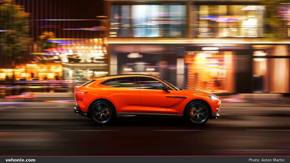 2026 Aston Martin DBX707 in orange, side profile with motion blur on a city street at night