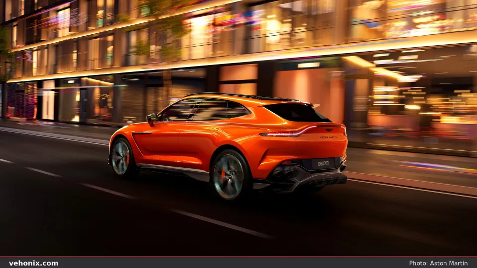2026 Aston Martin DBX707 in orange, rear three-quarter view on a city street at night