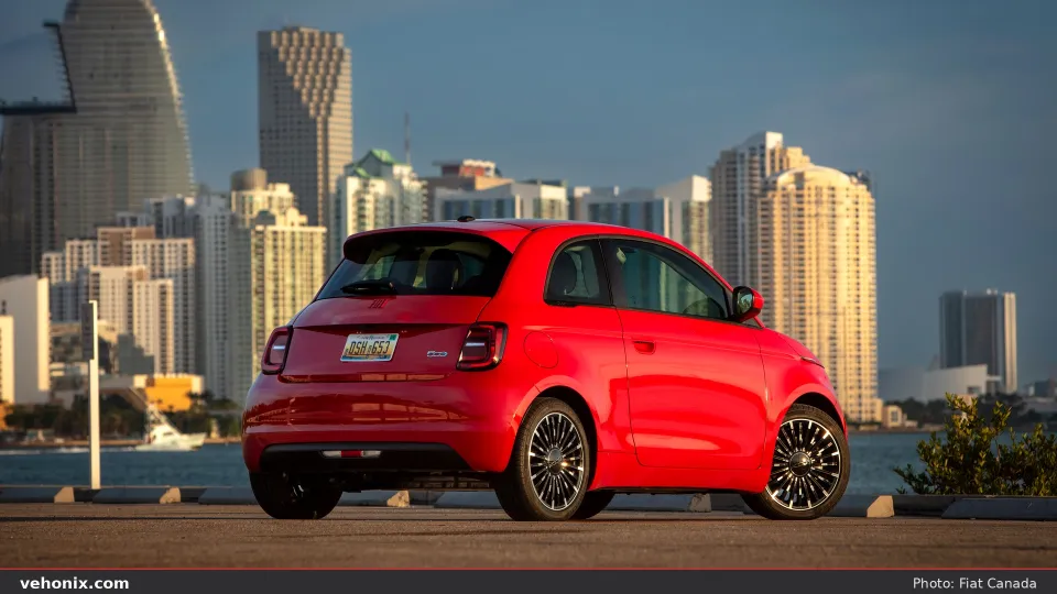 2026 Fiat 500e (RED) in red, rear three-quarter by a waterfront city skyline