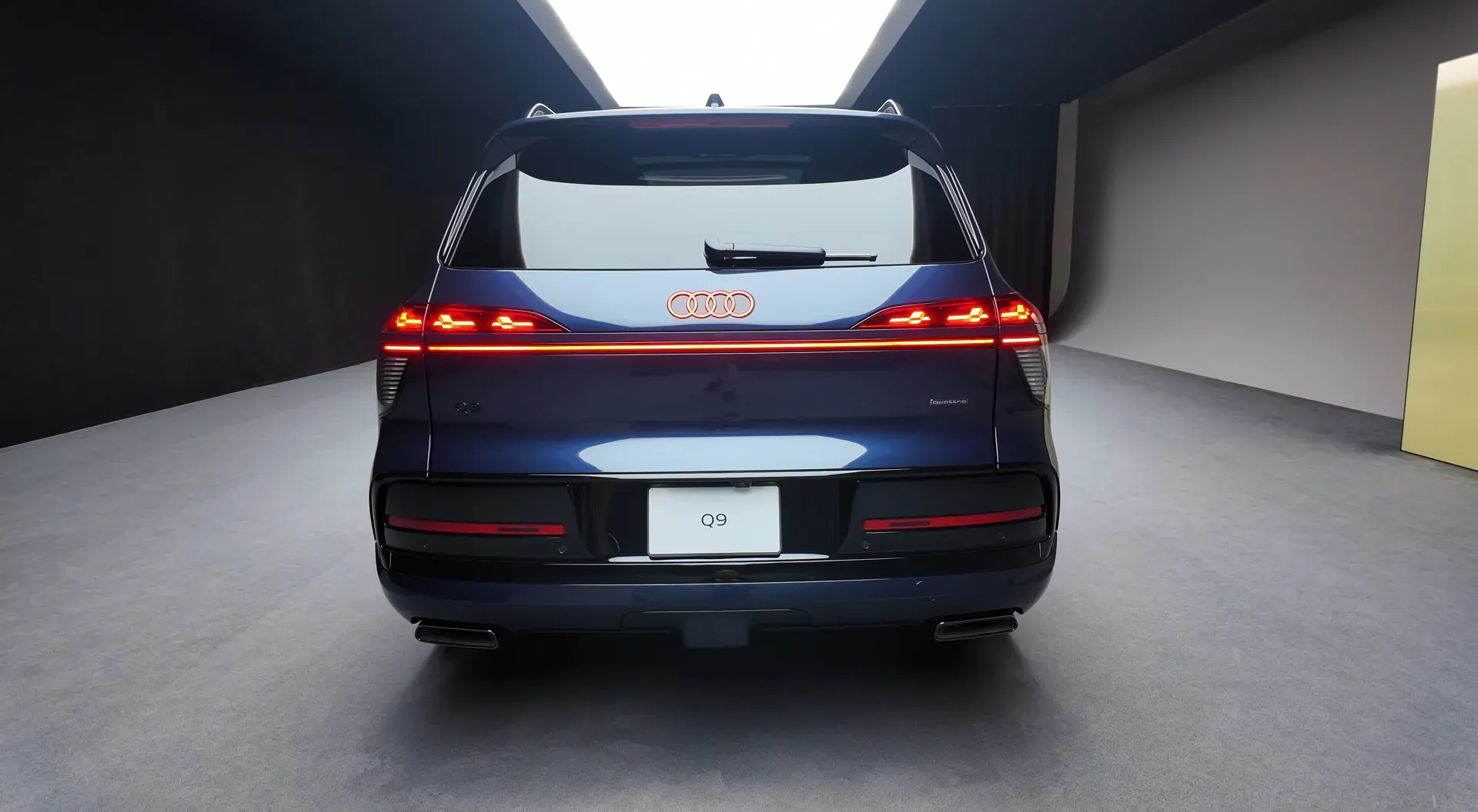 L'arrière d'un Audi Q9 2026 bleu foncé avec une barre de feux rouges sur toute la largeur et un logo Audi éclairé, dans un studio gris