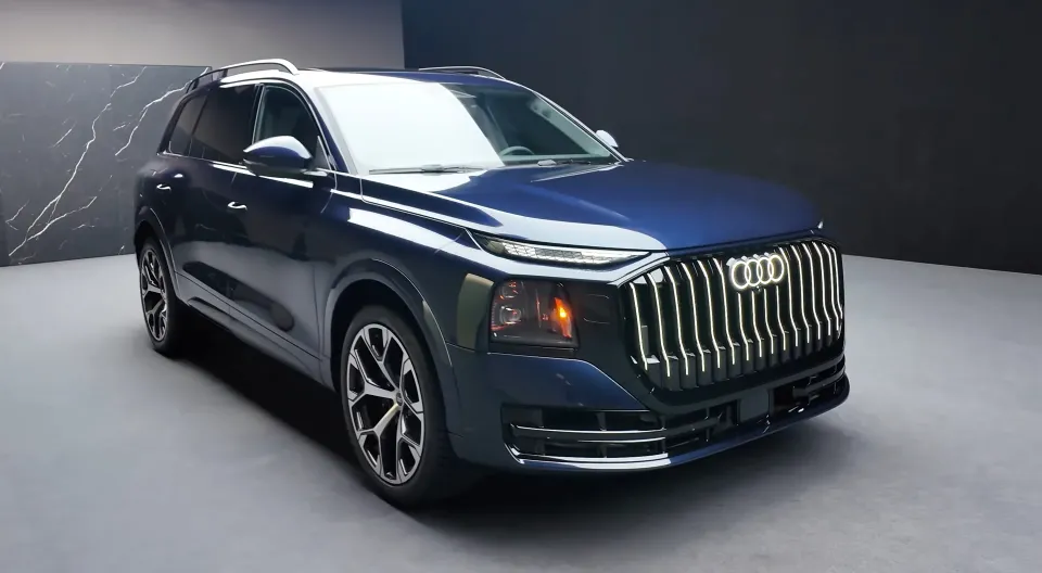 A dark blue 2026 Audi Q9 SUV with a lit vertical grille, seen from the front right in a grey studio
