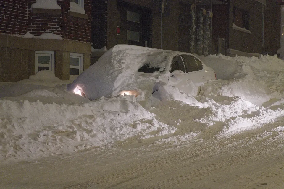 Une auto presque ensevelie sous une épaisse neige dans une rue la nuit, avec seulement le toit, les vitres et les phares allumés visibles