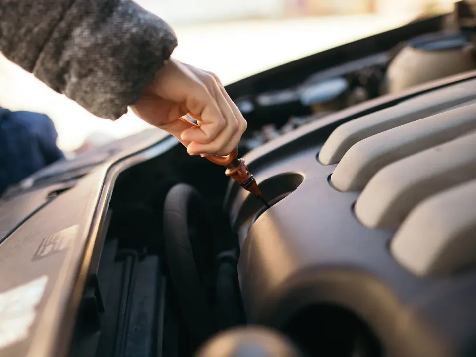 A person in a grey coat pulls an orange engine oil dipstick from under a car hood