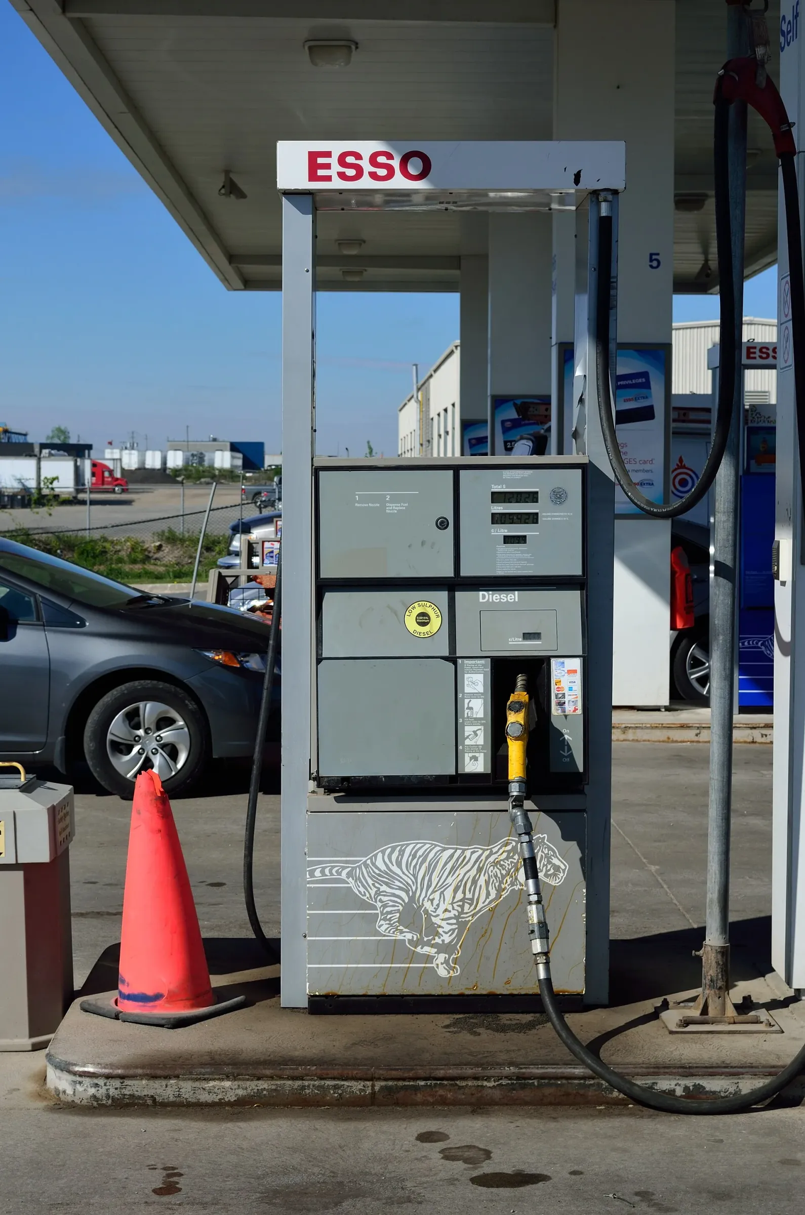 Une pompe à diesel Esso grise avec un pistolet jaune, un cône orange et une auto grise garée derrière, sous l'auvent d'une station-service