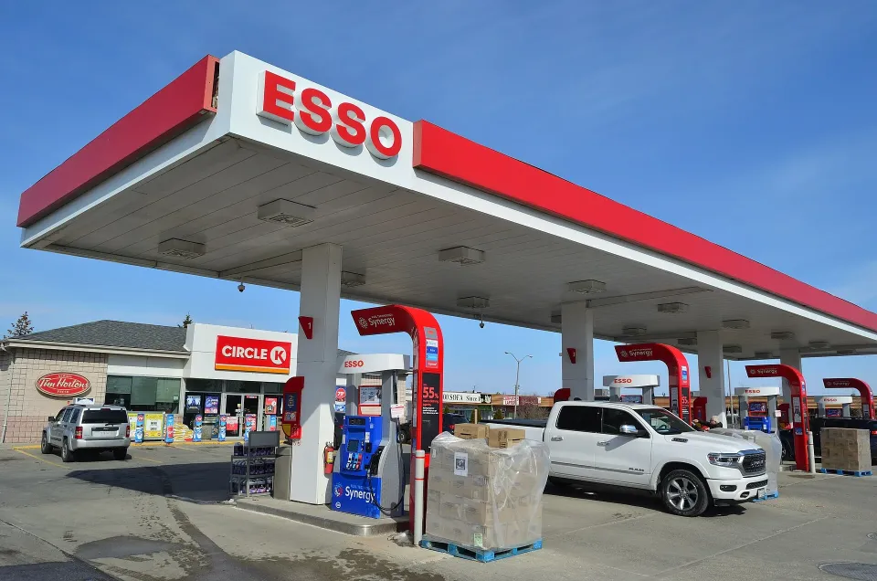 Une camionnette blanche garée aux pompes d'une station Esso avec un auvent rouge et blanc et un dépanneur Circle K par une journée ensoleillée