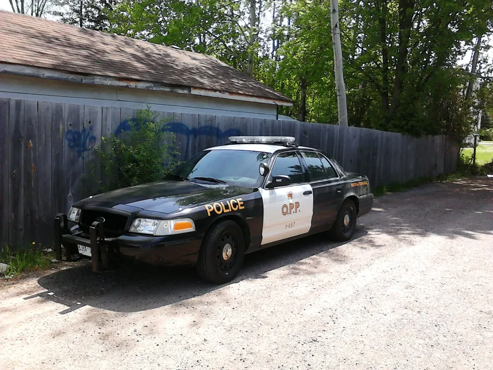 A black and white Ontario Provincial Police cruiser parked on gravel beside a wooden fence
