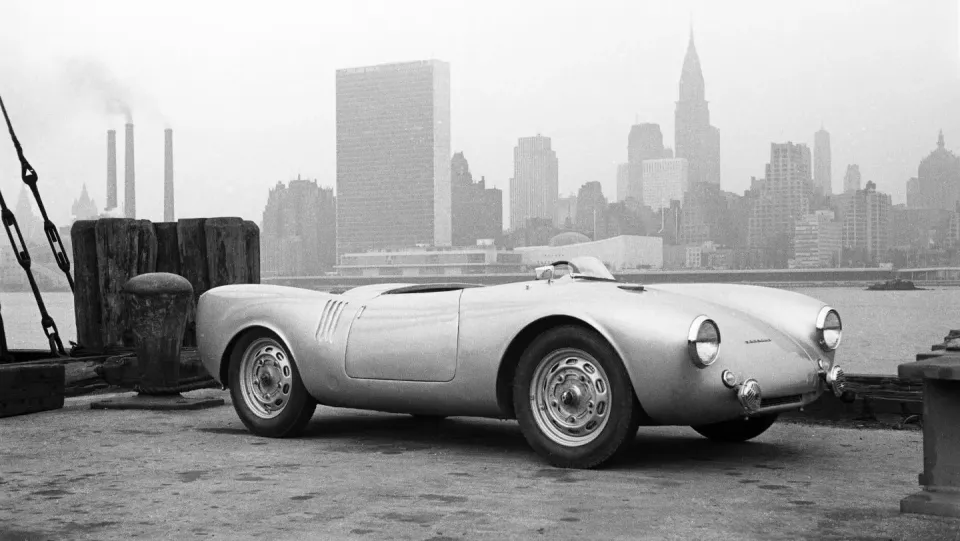 A black and white photo of a Porsche 550 Spyder on a pier with the New York skyline behind it