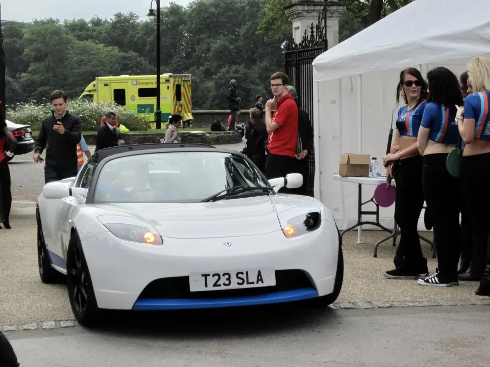 A white first-generation Tesla Roadster parked near a tent at an outdoor event