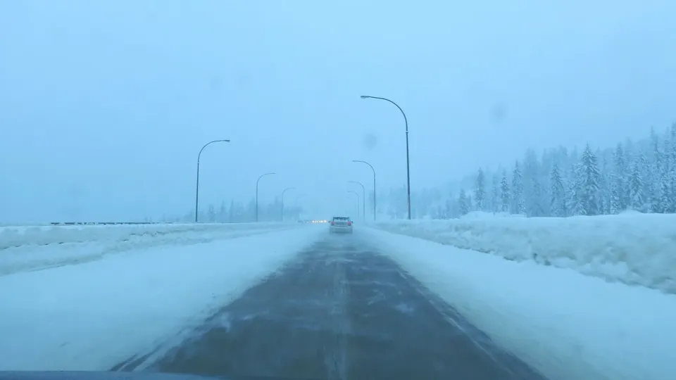 L'autoroute Coquihalla enneigée en Colombie-Britannique vue par le pare-brise, avec une auto devant, des lampadaires et des arbres sous la neige